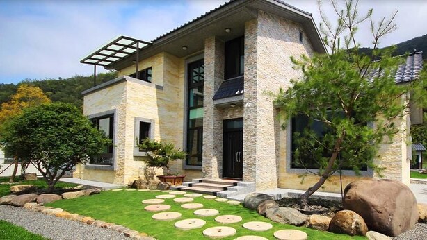 Una casa moderna de dos pisos con fachada de piedra, techo plano y un jardín cuidado con piedras circulares.
