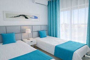 A hotel room with two beds, a nightstand with a lamp, a window with curtains, and a wall-mounted artwork. - T2-2º vista mar, piscina, bbq, para a praia vai a (Albufeira)