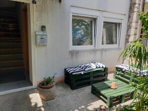 Un pequeño espacio al aire libre con un banco verde y una mesa de madera, una planta en maceta y una ventana con vistas a árboles.
