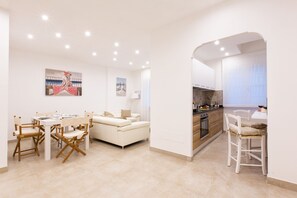 Una cocina moderna con armarios blancos, una zona de comedor con una mesa y sillas blancas, y un área de estar con un sofá y un cuadro en la pared.
