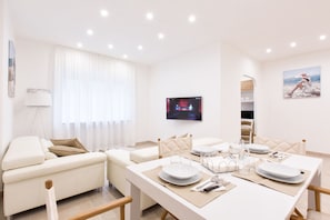 A modern living room with a dining area, white furniture, a flat-screen TV, and a painting on the wall.