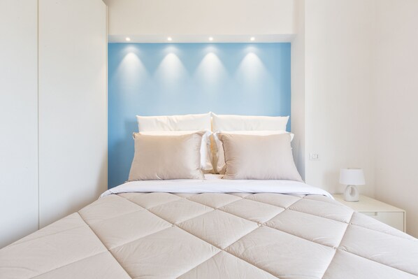 A neatly made bed with a quilted duvet, a bedside table with a lamp, and a wall with recessed lighting.
