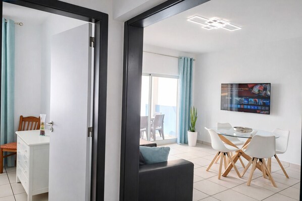 A modern living room with a dining area, a TV mounted on the wall, and a balcony visible through the door.