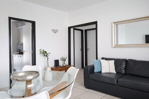 Un salon moderne avec une table à manger en verre, un canapé en cuir noir et une cuisine visible à travers une porte ouverte.
