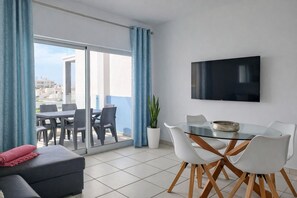 Un salon moderne doté d’une porte-fenêtre coulissante donnant sur un balcon, d’un téléviseur à écran plat fixé au mur et d’un coin repas avec une table en verre et des chaises blanches.