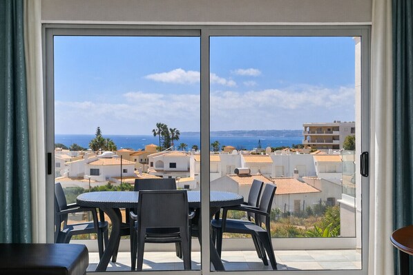 Varanda com mesa e cadeiras, porta de vidro deslizante e vista para edifícios e mar.
