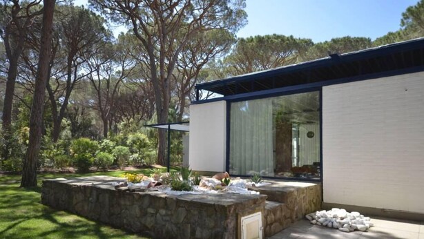 Una casa moderna con un muro di sostegno in pietra, ampie finestre e un giardino con alberi e arbusti.