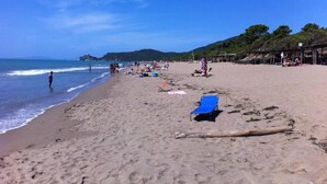 Una spiaggia con persone che camminano e siedono sulla sabbia, alcune sedie a sdraio e ombrelloni.
