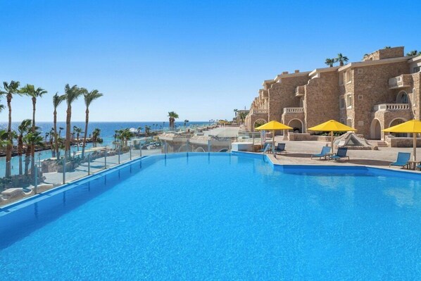 A pool area with clear blue water, surrounded by palm trees and a stone building with balconies. - Hotel Albatros Citadel (Sahl Hasheeh)