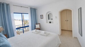 Una habitació d'hotel amb un llit, una cadira, una taula petita, una finestra amb cortines, una porta i vista al mar.