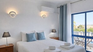 Un dormitori amb un llit, dues làmpades de nit, una finestra amb cortines i un balcó amb cadires.