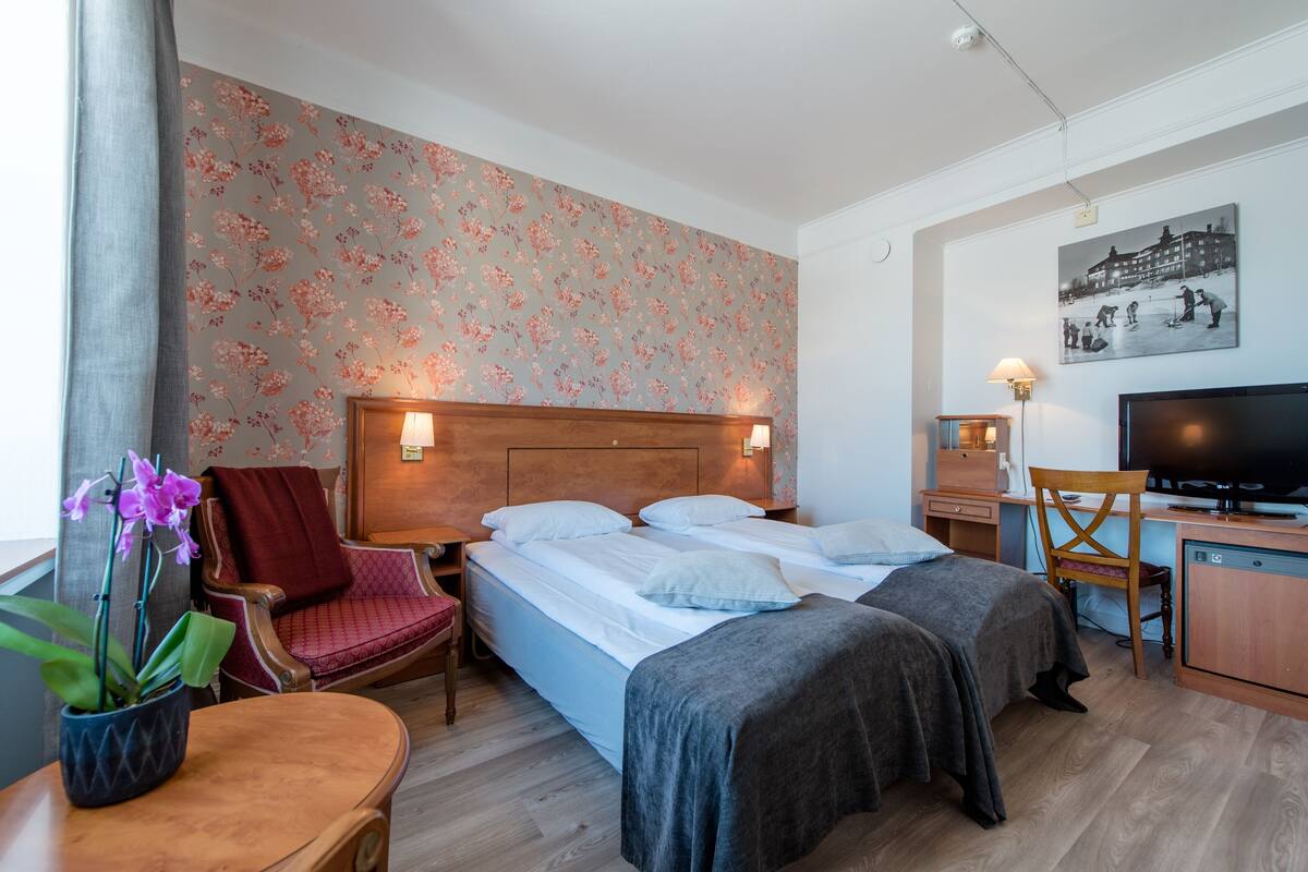 A hotel room with two beds, a desk, a TV, a chair, and a potted plant.