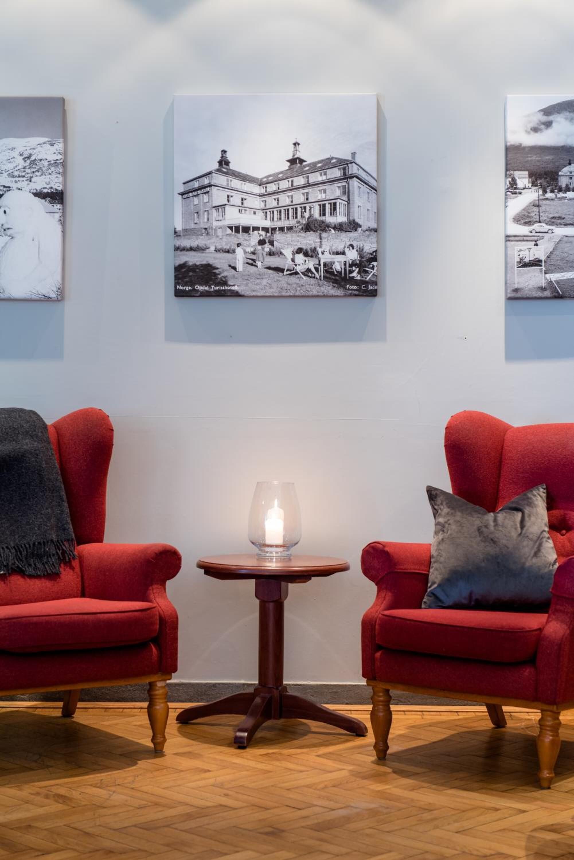 Una habitación con dos sillones rojos, una pequeña mesa de madera con una vela, y tres fotografías enmarcadas en la pared.