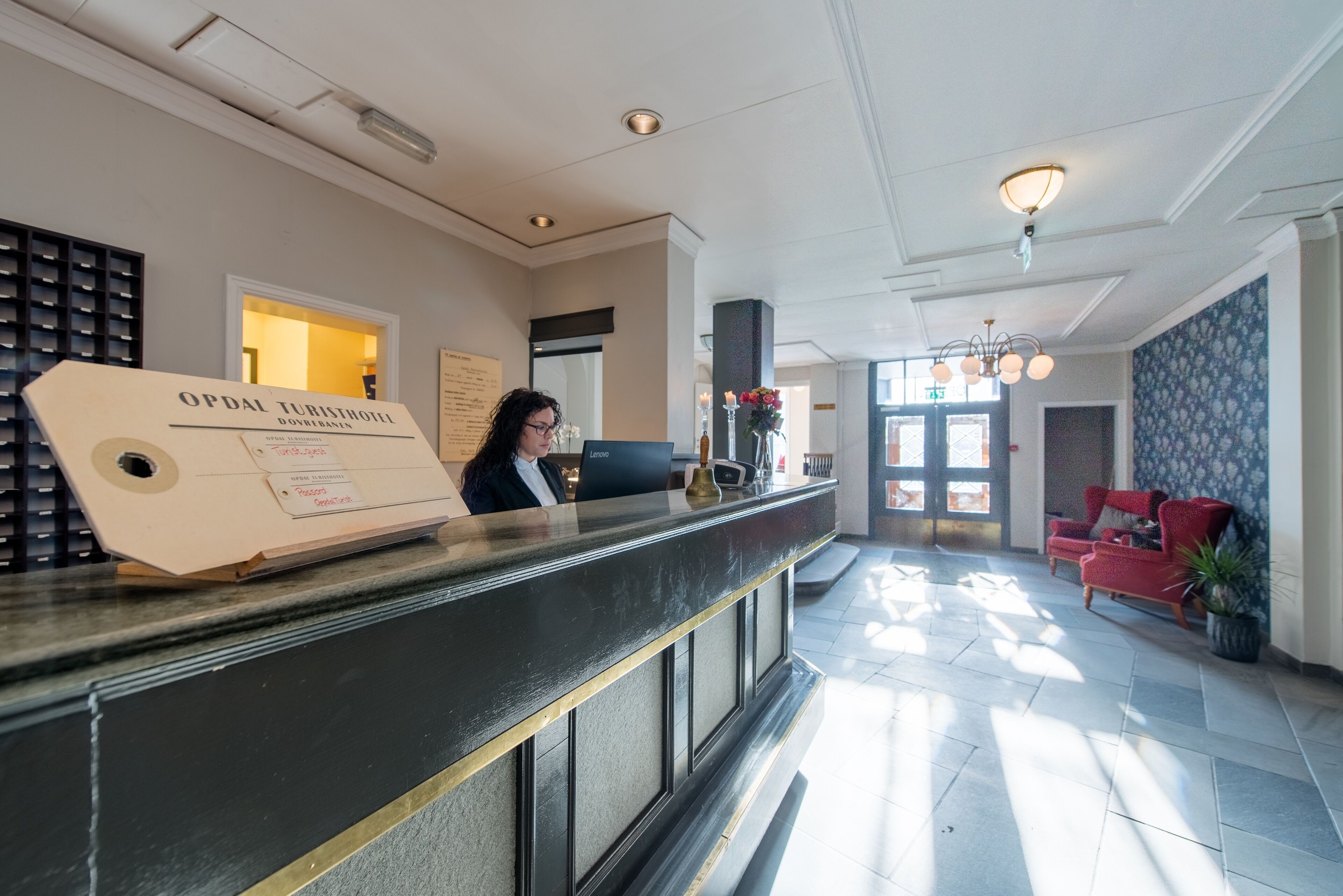 Zona de recepción de hotel con un mostrador, una recepcionista, un área de asientos con sillas rojas y un amplio ventanal que deja entrar luz natural.
