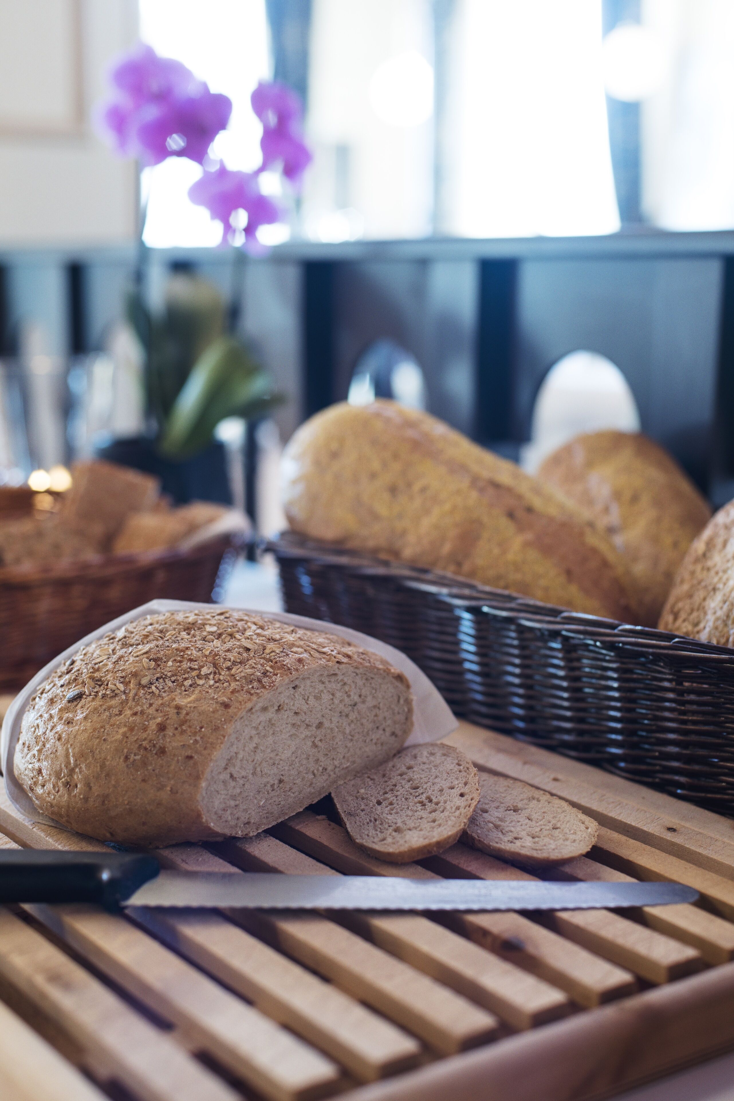 A loaf of bread on a cutting board with a knife, a basket of bread in the background, and a vase with purple flowers.