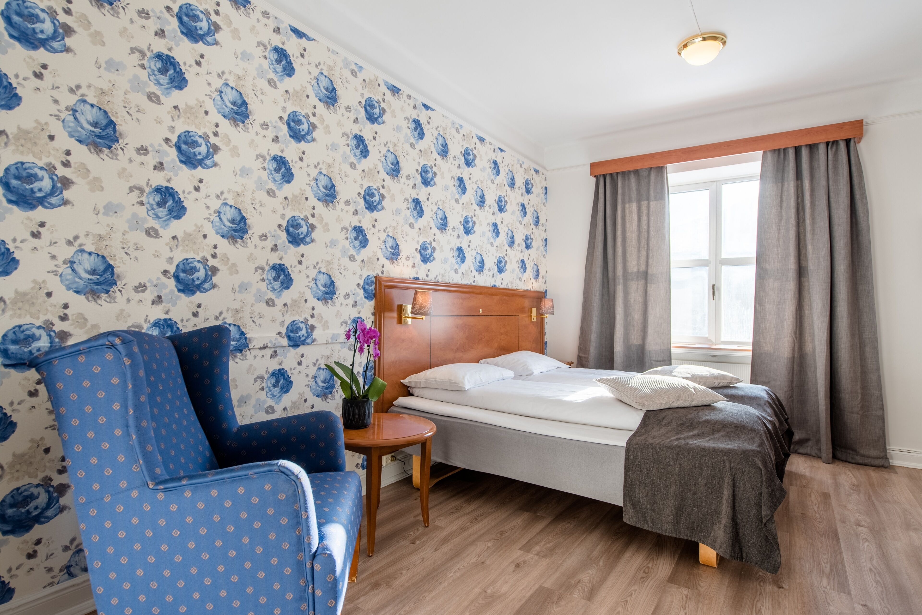 A bedroom with a wooden headboard, a single bed, a blue armchair, a small table with a potted plant, and a window with curtains.