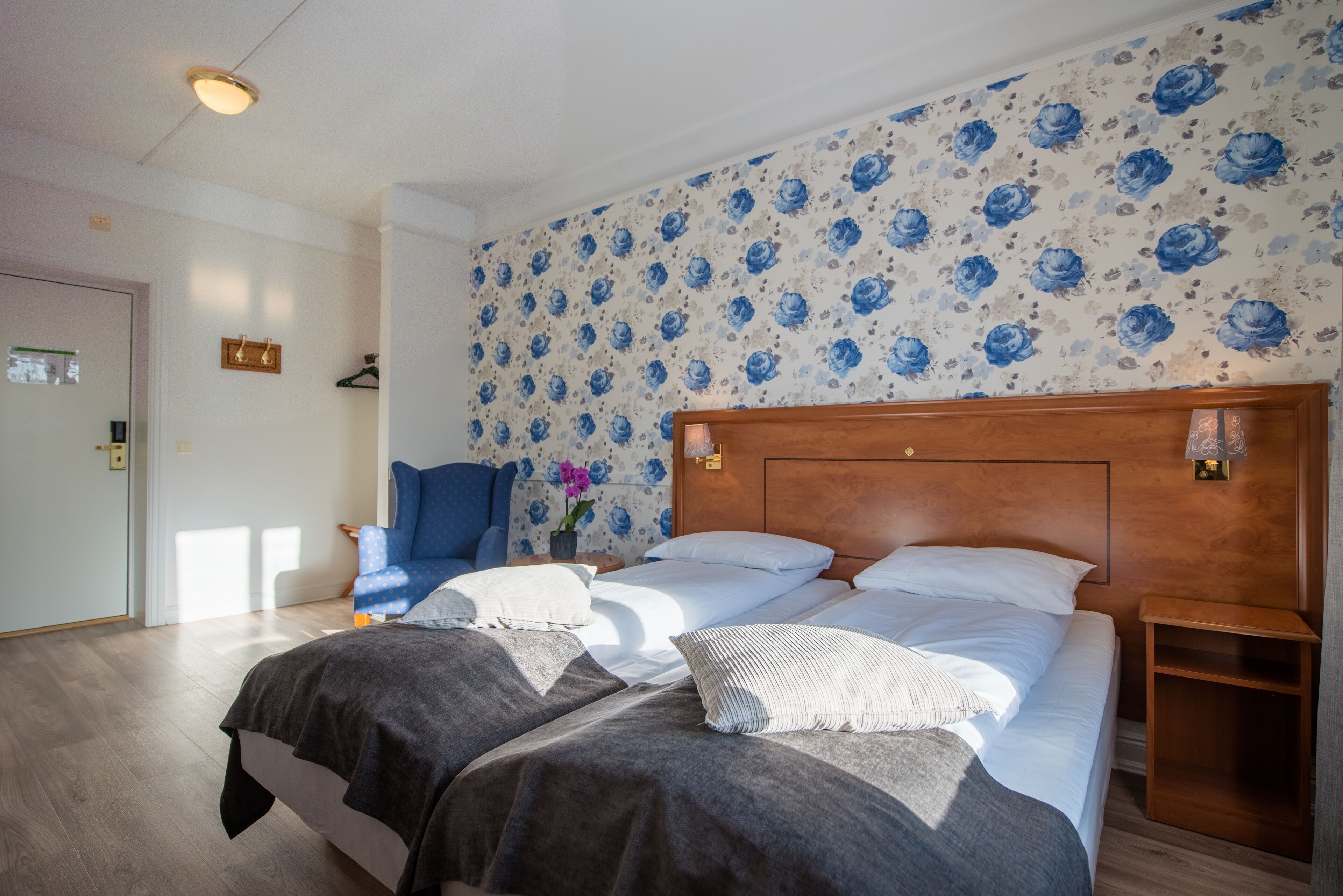 Ein Hotelzimmer mit zwei Betten, einem Holz-Kopfteil, floralem Wandpapier und einem blauen Stuhl.