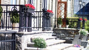Un edificio de piedra con una serie de escalones que conducen a una puerta, barandillas de metal negro y macetas con flores rojas.