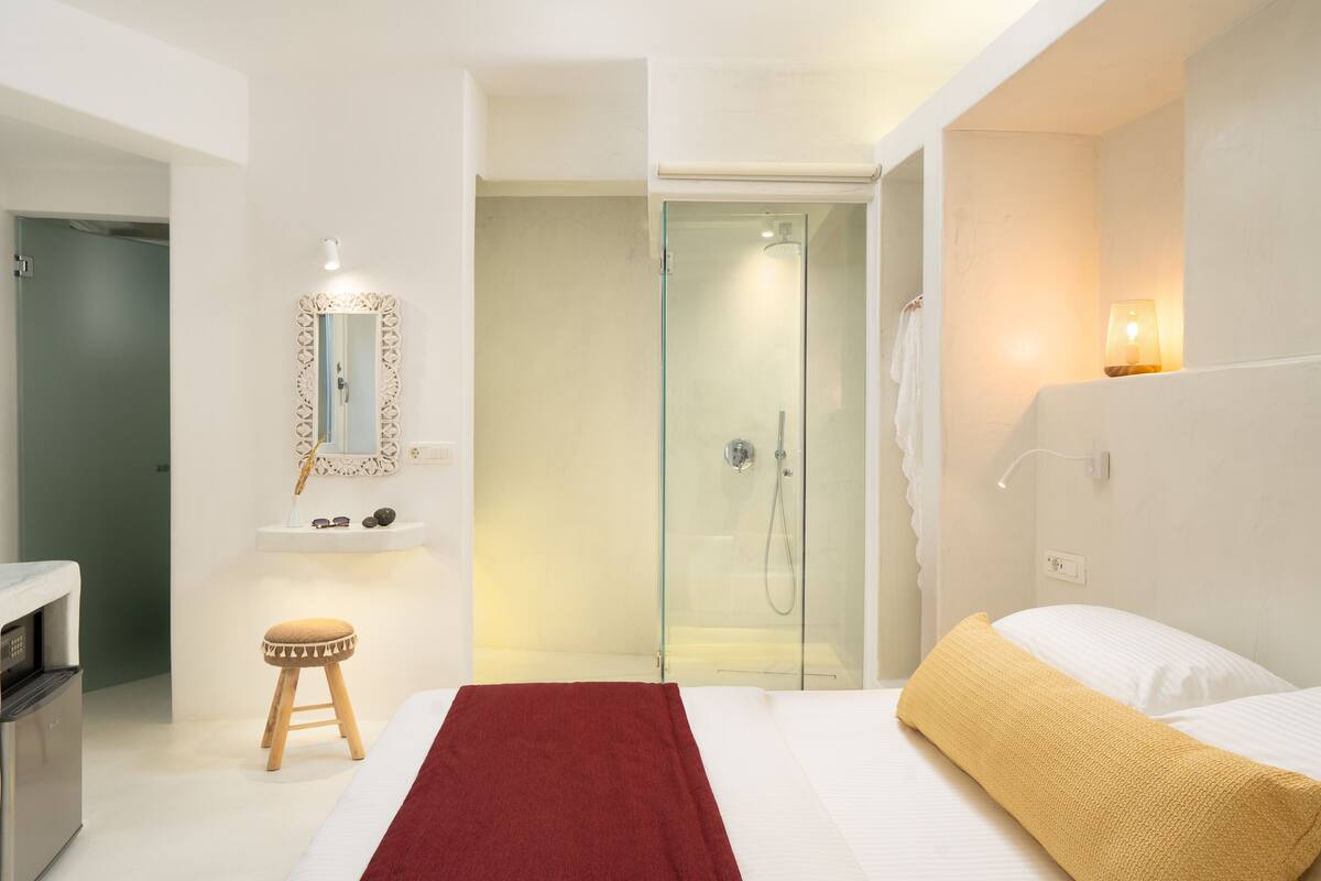 Una habitación de hotel con una cama, una toalla roja, un taburete de madera, una ducha con paredes de cristal y un espejo fijado a la pared.