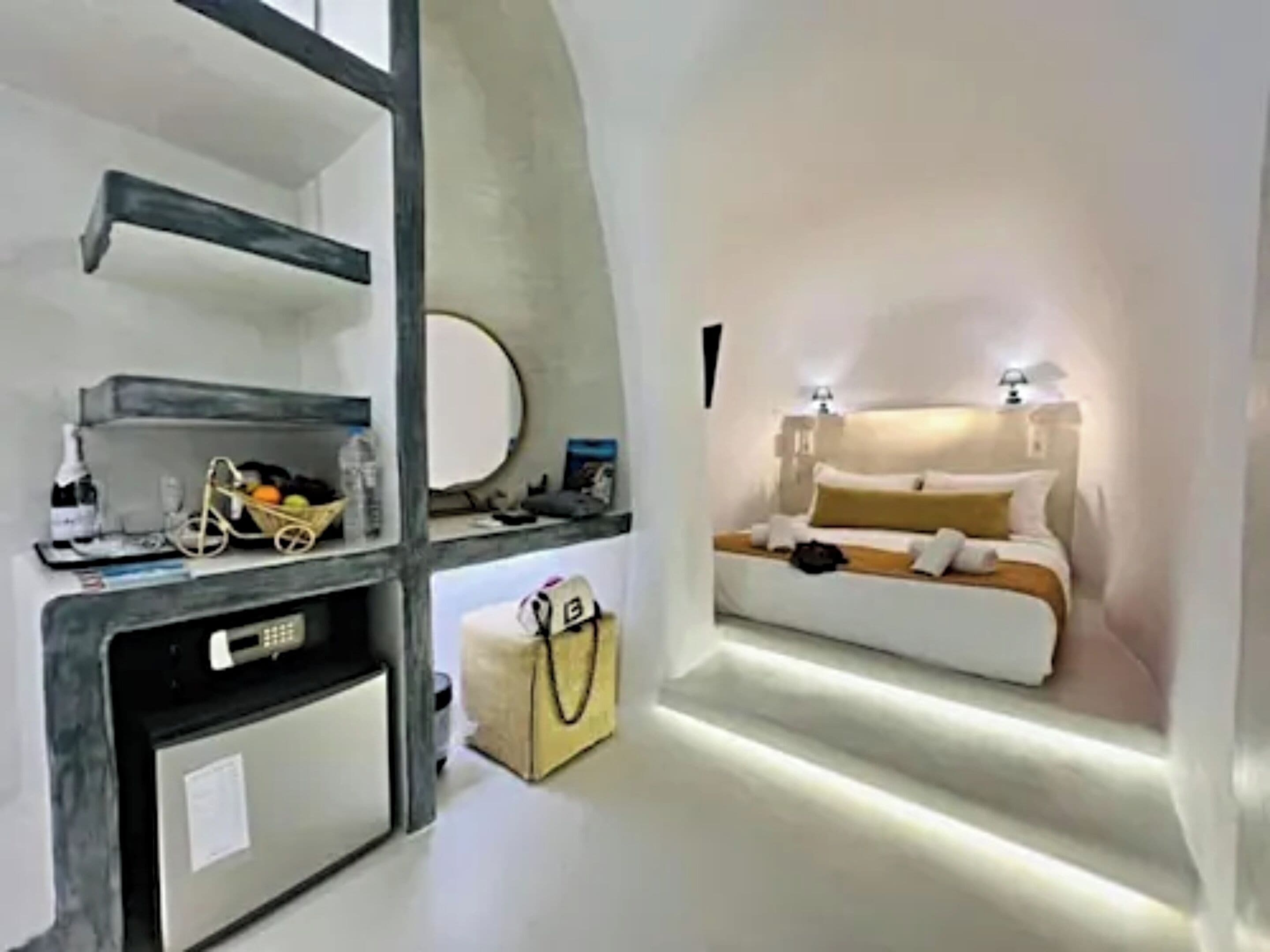 Une chambre d’hôtel avec un lit, des lampes de chevet, une petite table sur laquelle se trouvent une bouteille et un panier de fruits, une valise, et une étagère fixée au mur avec un miroir et un objet rond.