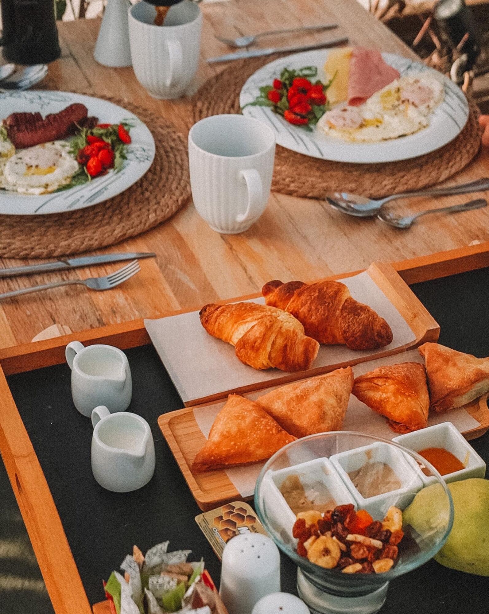 Un desayuno con croissants, bollería y un bol de fruta sobre una mesa de madera.