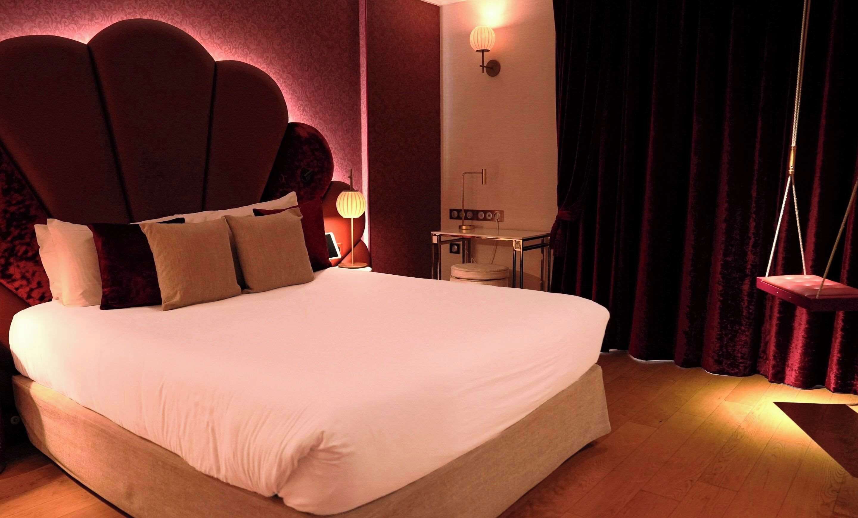 Una cama grande con ropa de cama blanca, cabecera con diseño festoneado, mesita de noche con lámpara y una hamaca colgante.