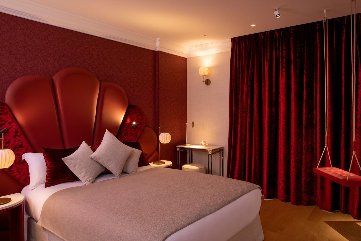A modern hotel room with a large bed, a red headboard, a bedside table with a lamp, and a red curtain.