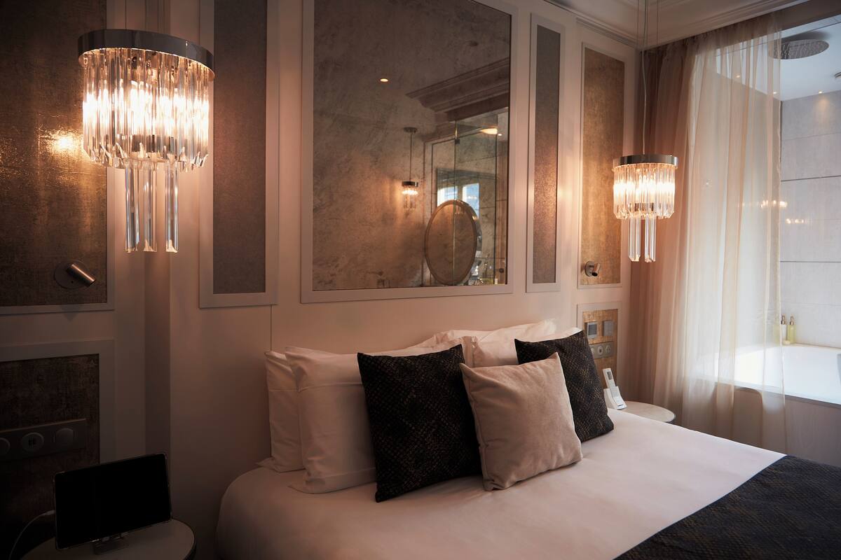 A modern hotel room with a large bed, two wall-mounted chandeliers, a large mirror, and a view of the cityscape.