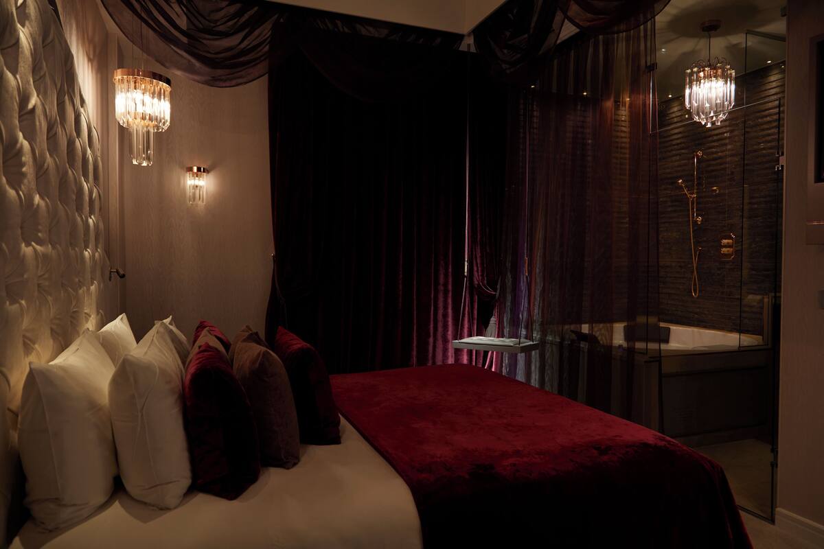 A bedroom with a bed, pillows, a red blanket, a glass shower, and two wall-mounted lights.