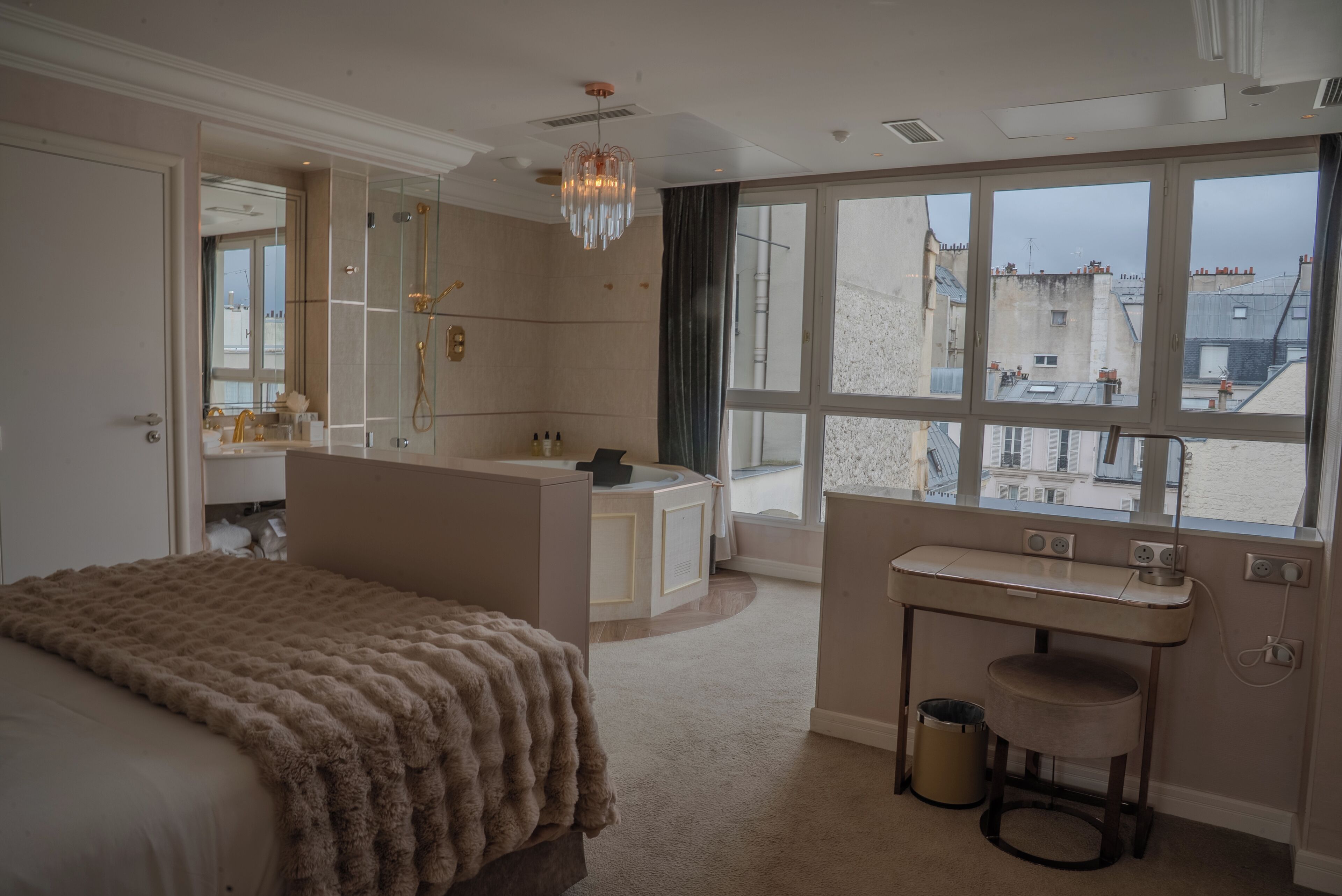 A hotel room with a large bed, a vanity with a mirror, a chandelier, and a view of buildings through large windows.