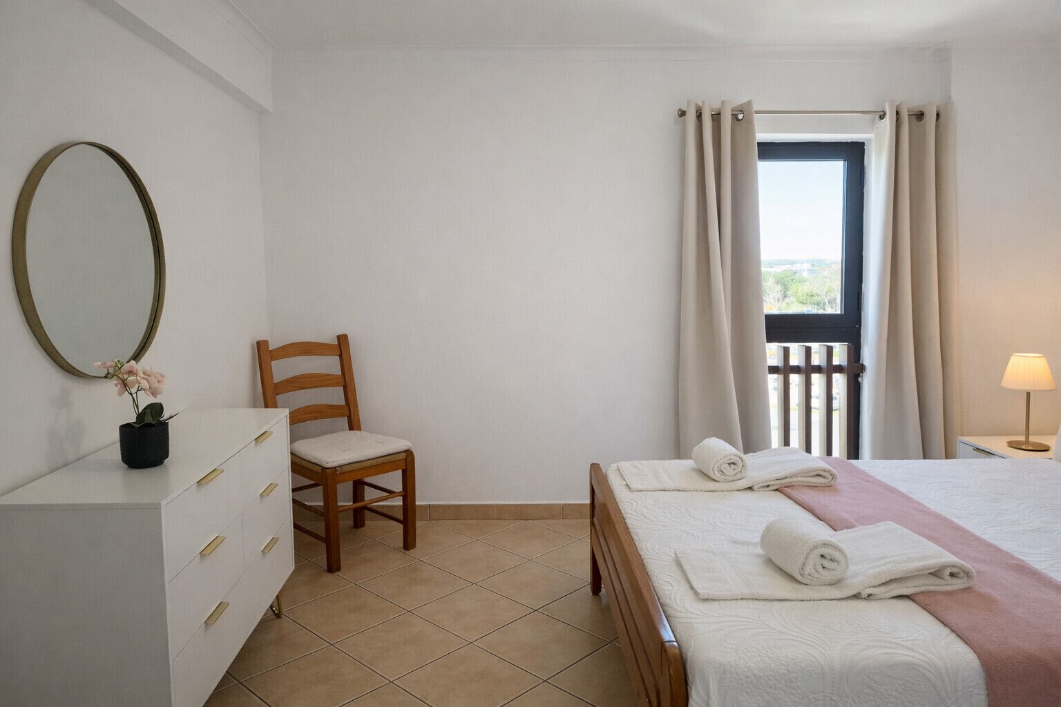 A bedroom with a bed, a chair, a dresser, a mirror, a window with curtains, and a lamp.