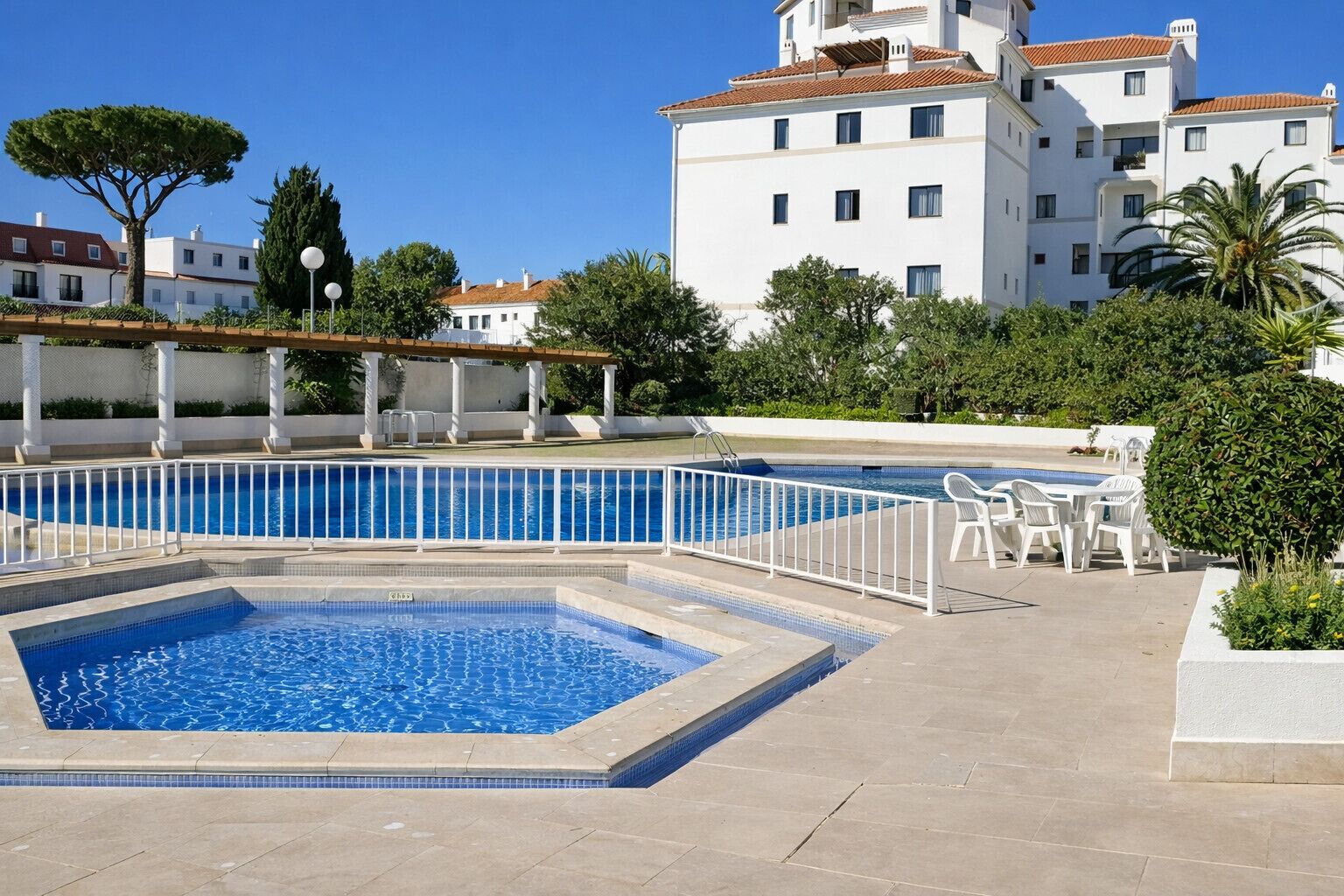 A swimming pool with a surrounding deck, outdoor seating, and a building with balconies in the background.