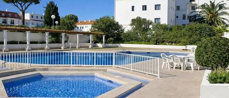 A swimming pool with a surrounding deck, outdoor seating, and a building with balconies in the background.