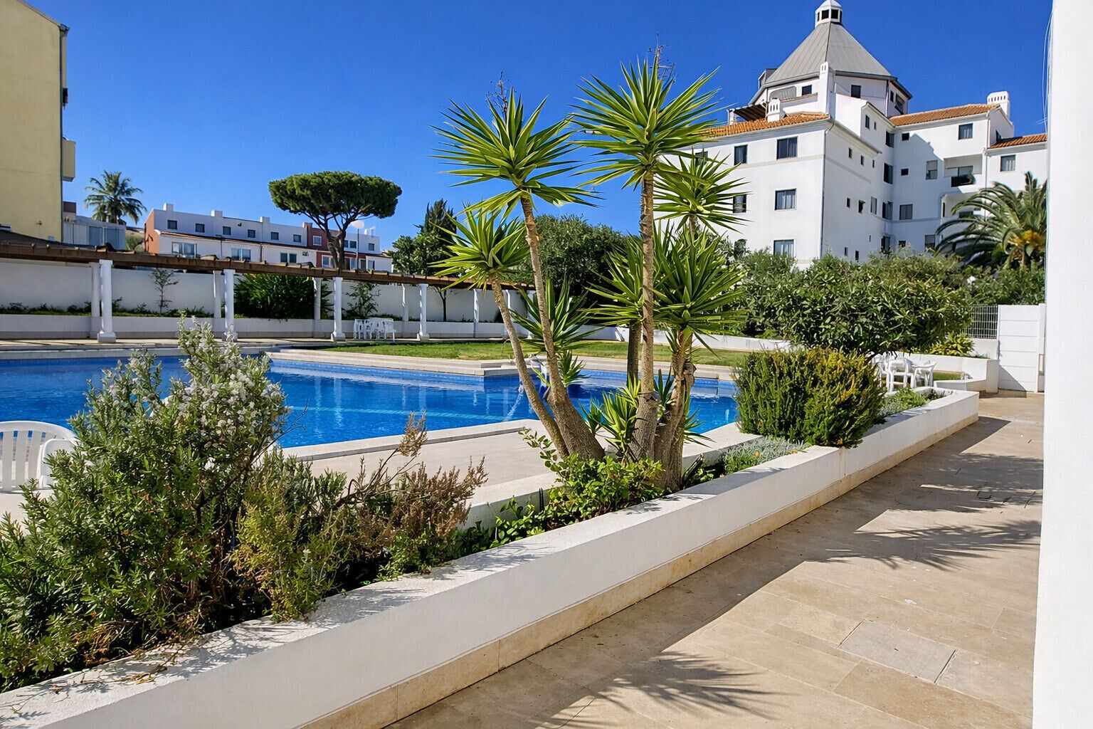 Ein von Grün umgebener Pool, ein weißes Gebäude mit Kuppel und ein klarer, blauer Himmel.