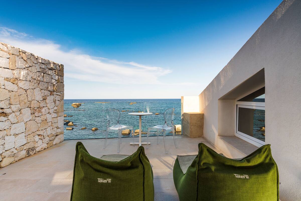 A balcony with a stone wall, two green chairs, a small table, and a view of the sea.