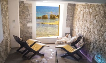 Una habitación con paredes de piedra, un ventanal con vistas al mar y dos tumbonas con cojines amarillos.