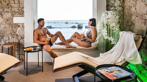 A room with a stone wall, a window overlooking the sea, a small table with fruits, and a lounge chair with a magazine. - Re Dionisio Luxury Suites (Syracuse)