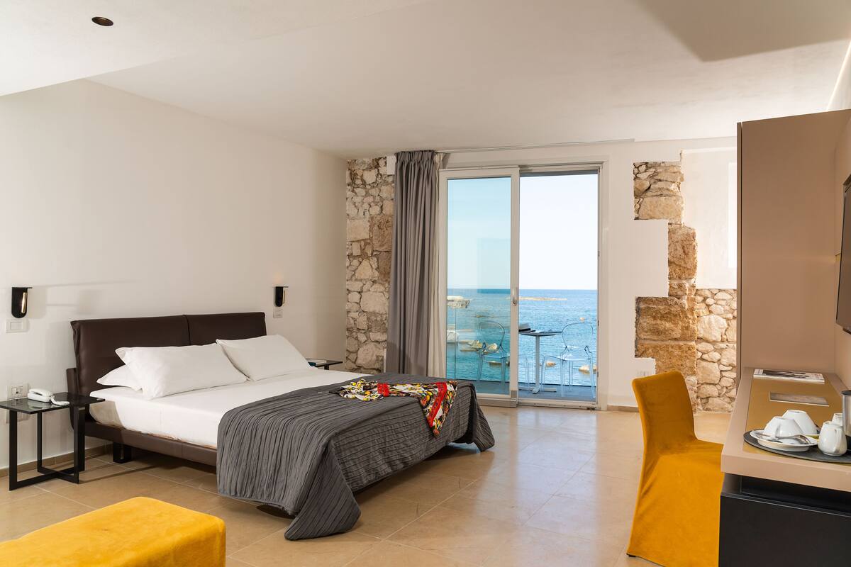 A hotel room with a bed, a desk, a chair, and a view of the sea.