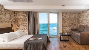 A bedroom with a bed, a sofa, a small table, and a view of the sea through a glass door. - Re Dionisio Luxury Suites (Syracuse)