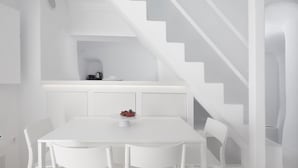 A modern, white interior with a dining area featuring a table and chairs, a kitchen area with a kettle, and a staircase. - Oia White Cave Oia Cliff (Santorini)