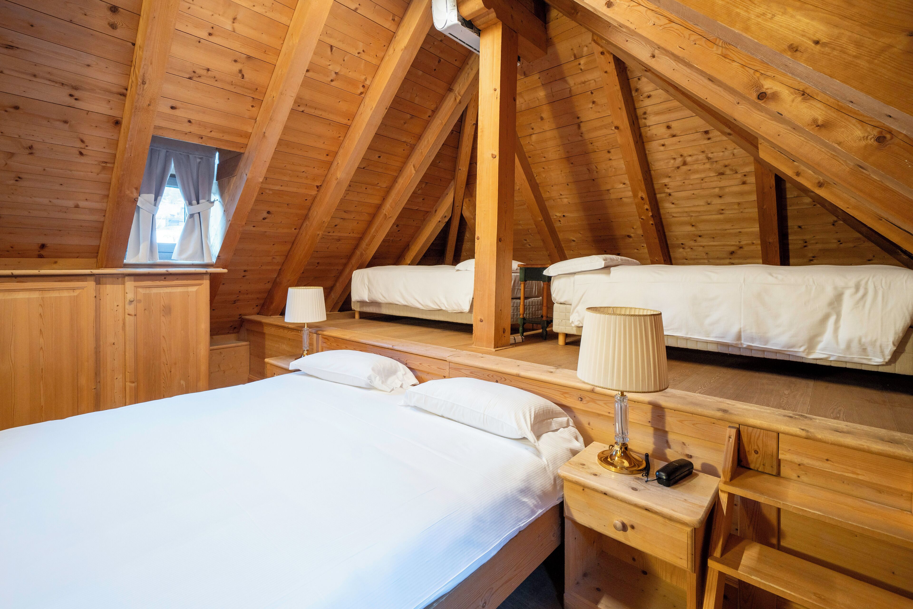 A wooden attic bedroom with two beds, a nightstand with a lamp, and a window with curtains.