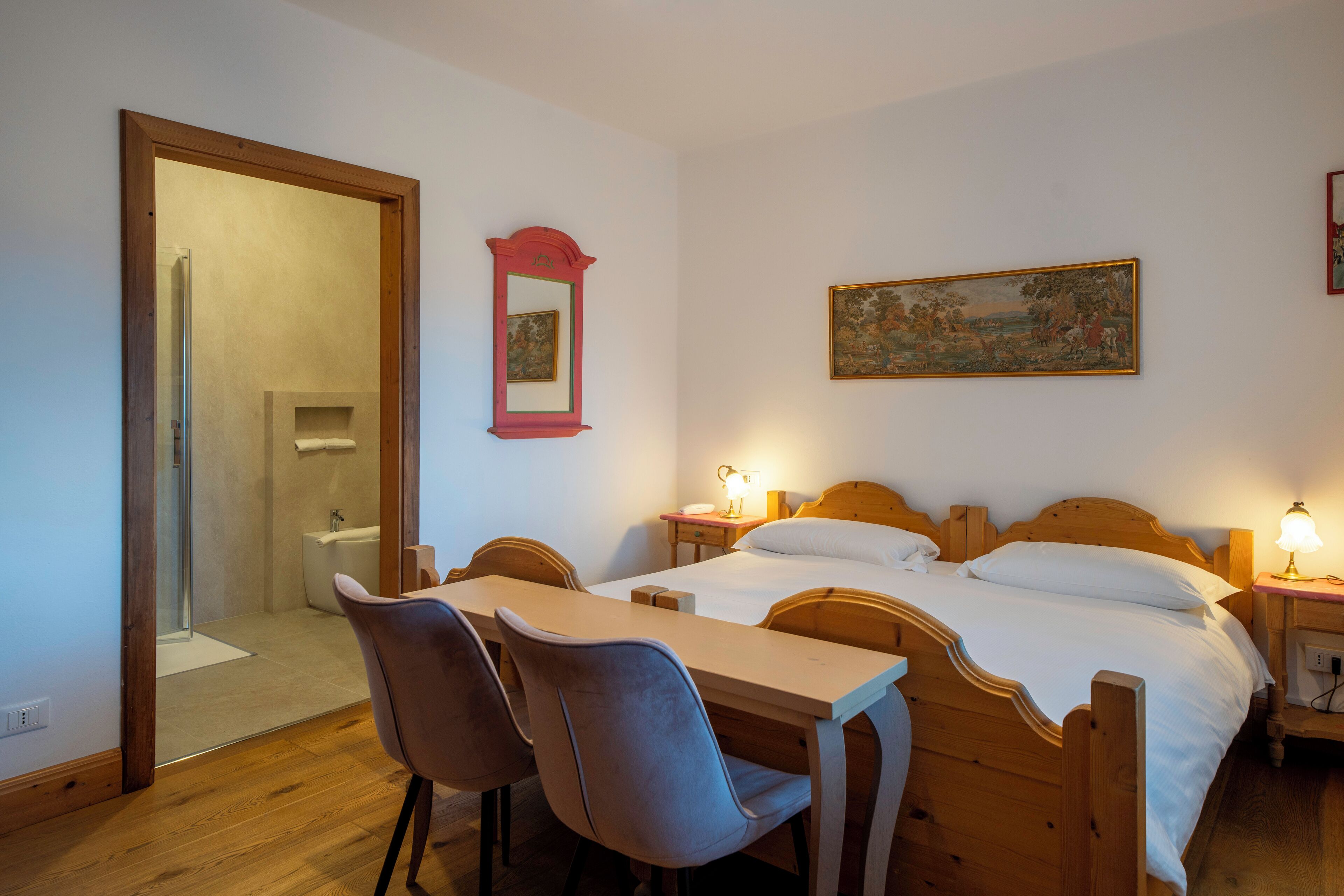 A bedroom with a wooden bed, a desk, two chairs, a small bedside table with a lamp, a mirror, and a painting on the wall.