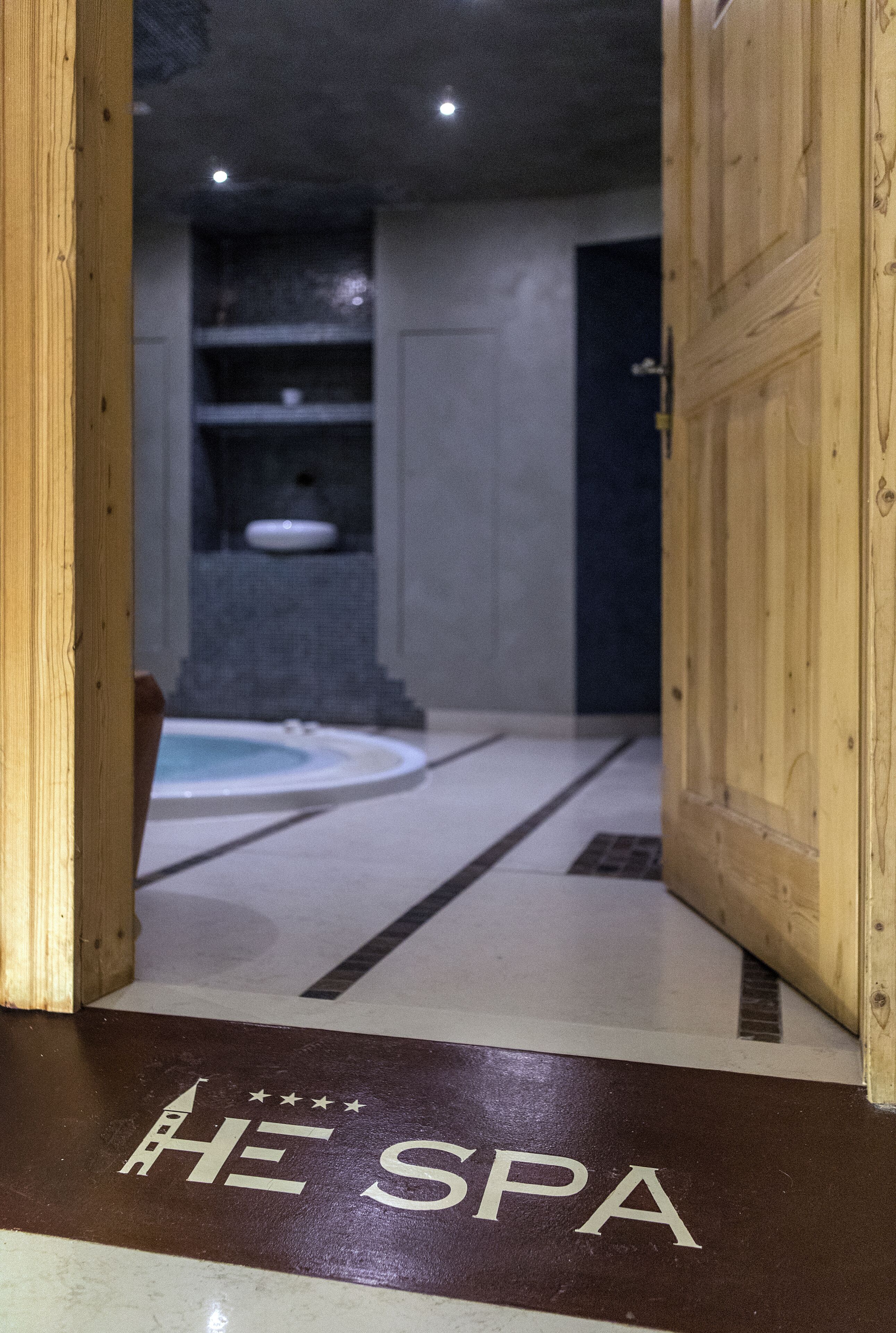 Entrada de um spa com porta de madeira, piso ladrilhado e placa indicando "HE SPA".
