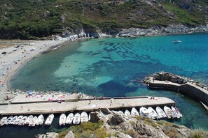 A coastal area with a clear blue sea, a sandy beach, and a rocky outcrop with boats docked.