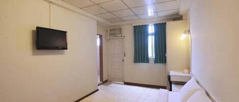 Habitación de hotel con cama, televisor montado en la pared, ventana con cortinas y mesita de noche con lámpara.