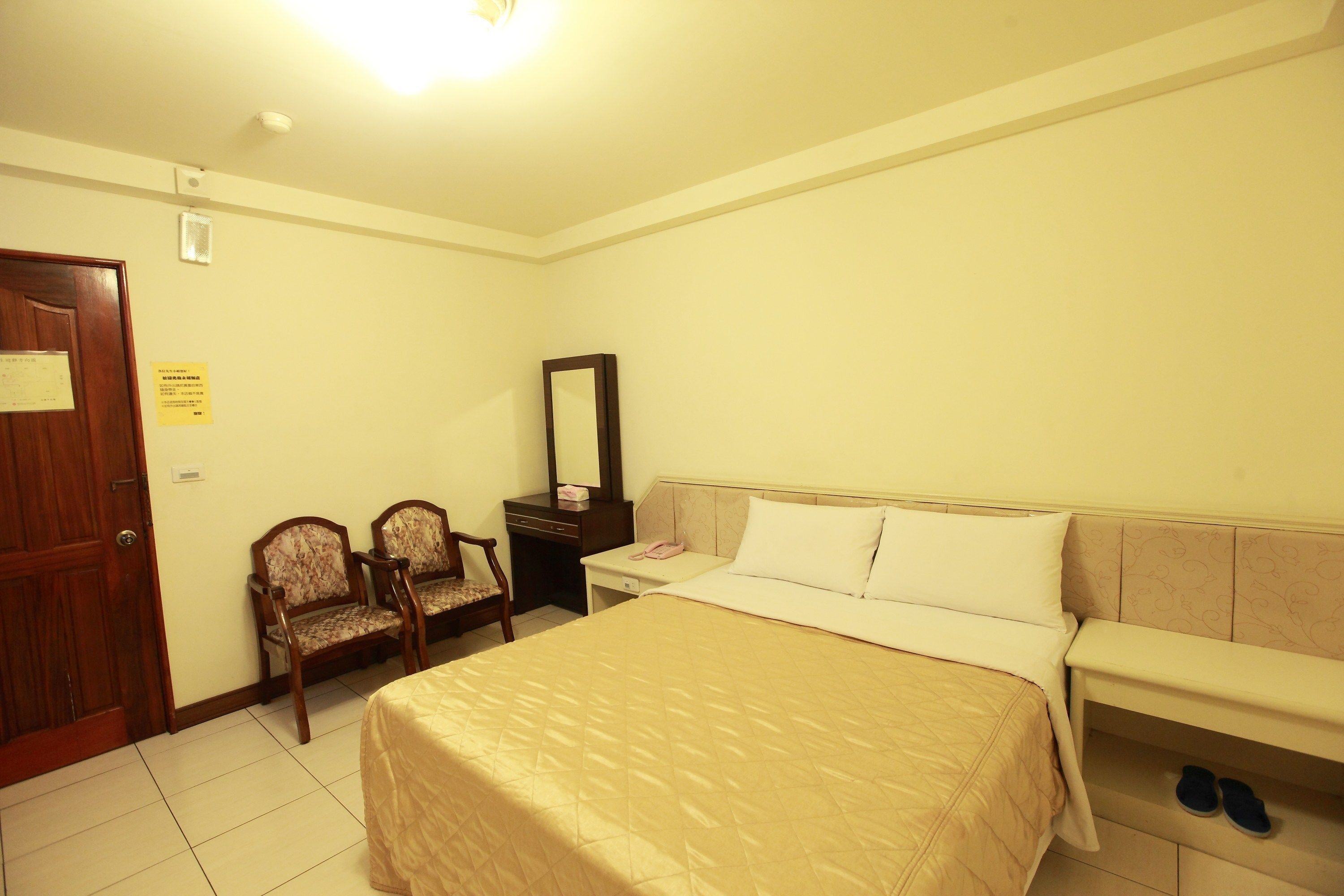 A hotel room with a bed, a desk, two chairs, a mirror, and a pair of slippers on the side table.