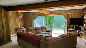 A cozy living room with a brown sofa, a coffee table, and a television. There is a wooden wall with a deer head decoration and a bookshelf with various items.