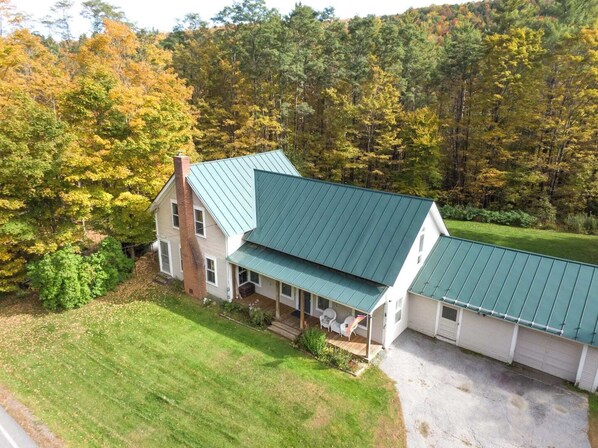 A two-story house with a green metal roof, surrounded by a well-maintained lawn and dense forest. - Classic Vermont Farmhouse near Sugarbush Resort (Warren)