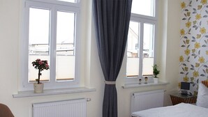 Un dormitorio con cama, dos ventanas con cortinas, un radiador, un escritorio con ordenador y una planta en maceta.