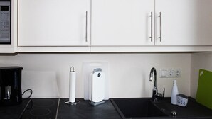 Una cocina con armarios blancos, encimera negra, cafetera, dispensador de agua, lavamanos y tabla de cortar verde.